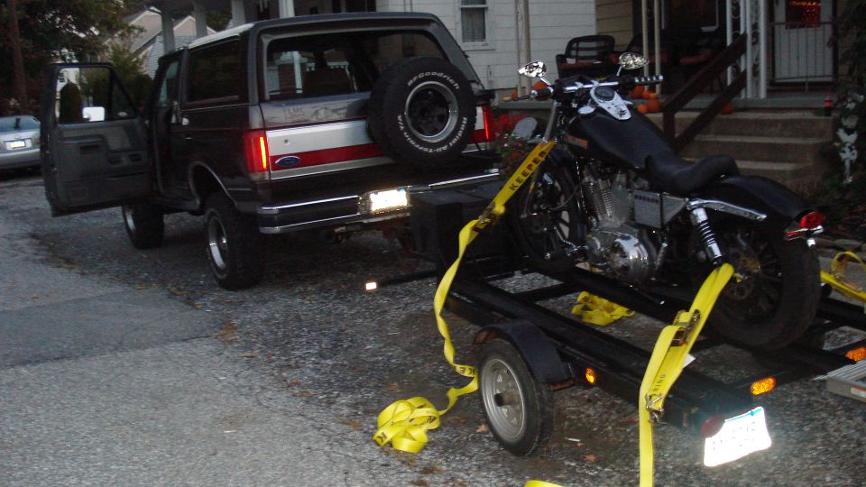 Ford Bronco with door open, towing a motorcycle on a trailer. License plate visible