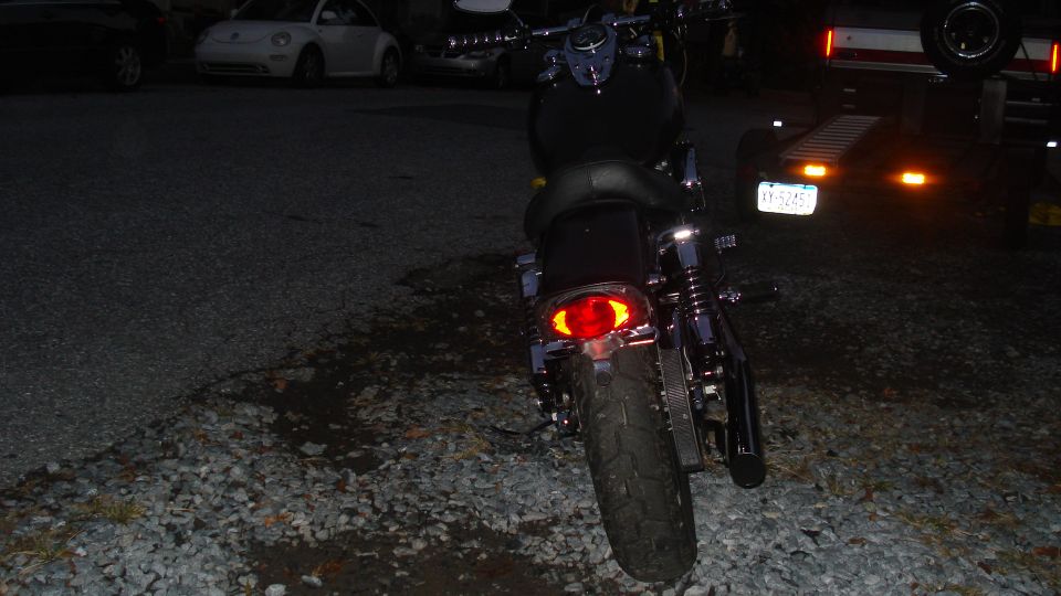 Motorcycle rear view, dark, gravel lot, trailer with lights in background