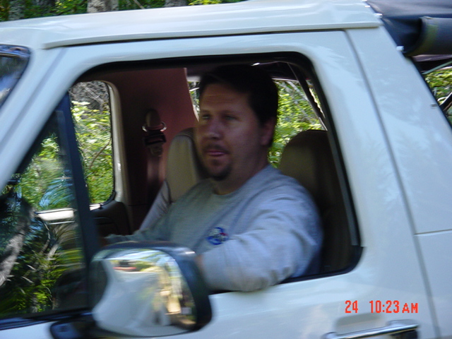 Man with goatee in gray sweatshirt driving a white SUV, arm on the door