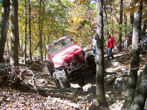 1992 Jeep YJ - A red Jeep Wrangler is shown off-roading on a challenging, rocky path through a forest with autumn foliage.