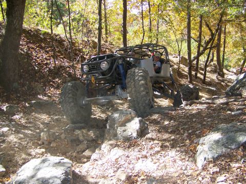 1992 Jeep YJ - A custom-built buggy with a roll cage and large tires is shown traversing a challenging rocky terrain in a wooded area.