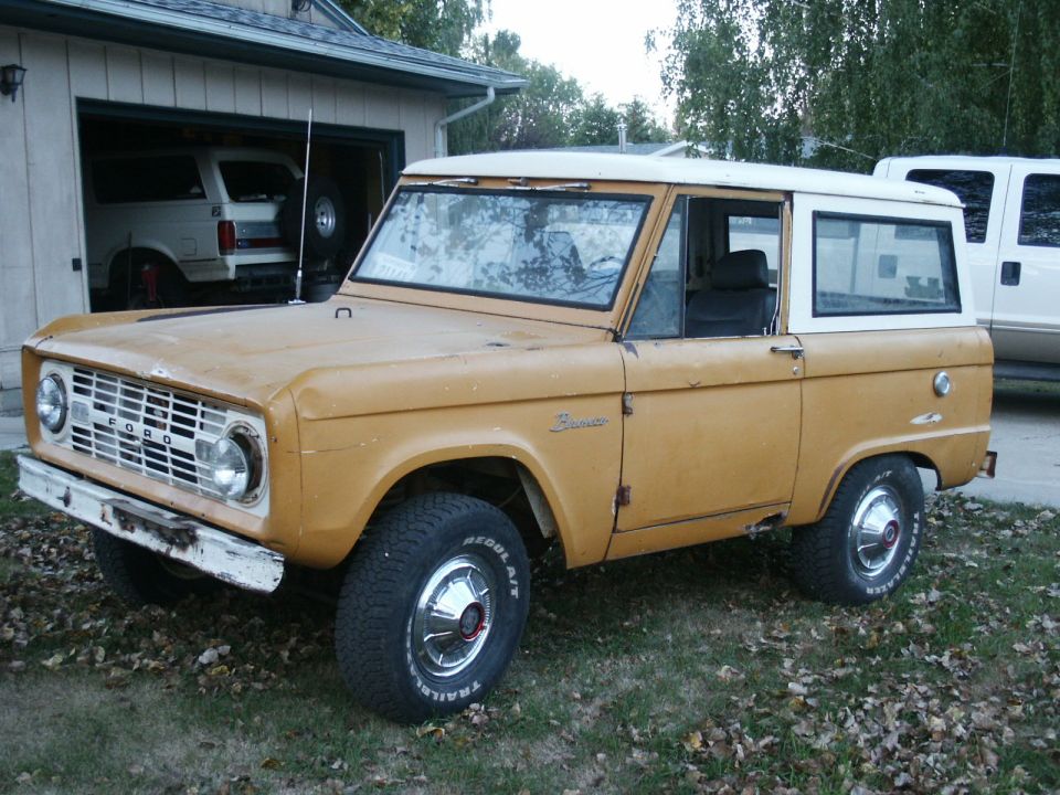 Yellow 1966 Ford Bronco with a white hardtop, parked in front of a garage. The vehicle is slightly weathered