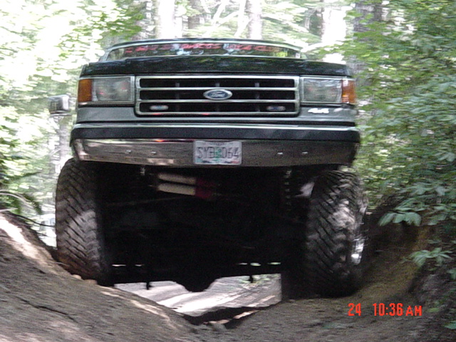Lifted 1987 Ford Bronco front view, driving on a dirt trail with trees in background. License plate SYB-064
