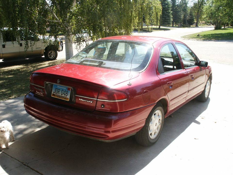 Red Mercury Mystique sedan, rear three-quarter view on driveway with sign in rear window