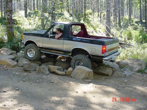 1989 Ford Bronco - A two-tone Ford Bronco is driven over large rocks on a dirt path surrounded by trees.