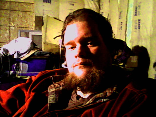 Bearded man in red jacket seated in a cluttered dim room.