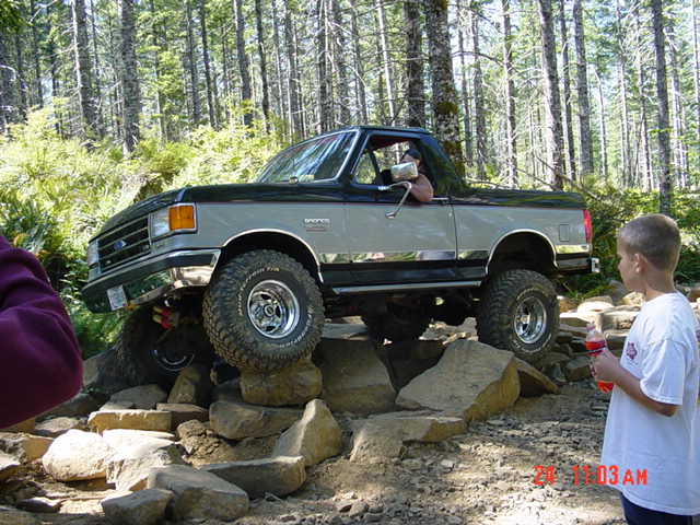 Green and white Ford Bronco climbing a rock pile on a forest trail