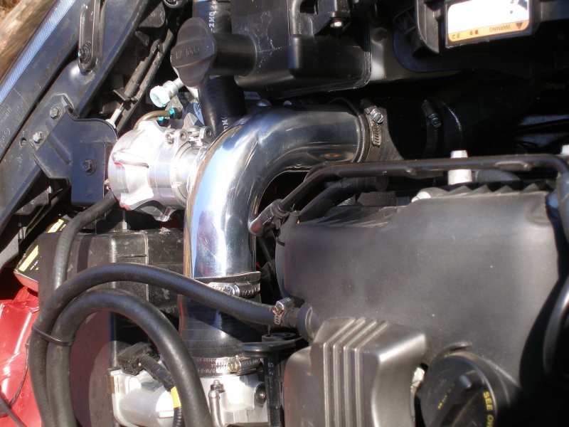 Engine bay showing chrome intake piping connected to a silver component, with black hoses and engine covers visible