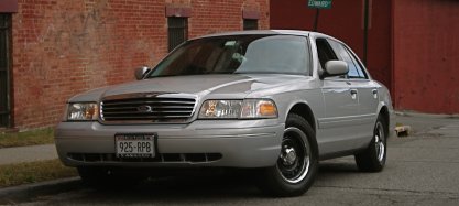 2000 Ford Crown Victoria - A silver 2000 Ford Crown Victoria sedan is parked on a street with brick buildings in the background.