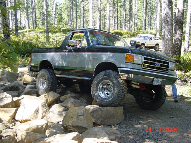 Lifted green and silver Ford Bronco topless, crawling a rock pile on a forest trail