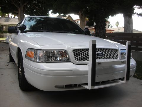 2004 Ford Crown Victoria - Three-quarter front view of a white Ford Crown Victoria sedan parked on a driveway under trees, fitted with a white police-style push bumpe…
