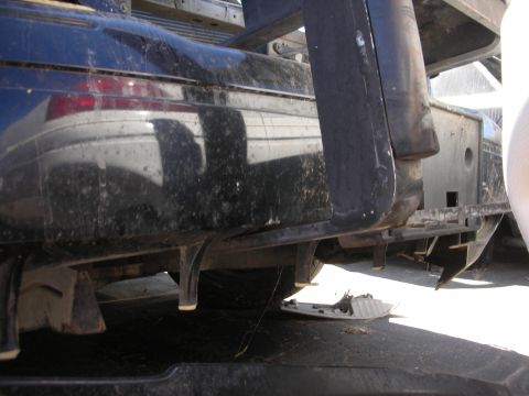 2004 Ford Crown Victoria - Low-angle view under the rear of a dark blue car resting on a truck or trailer deck, with a dirt-speckled rear bumper cover, red tail lamp…