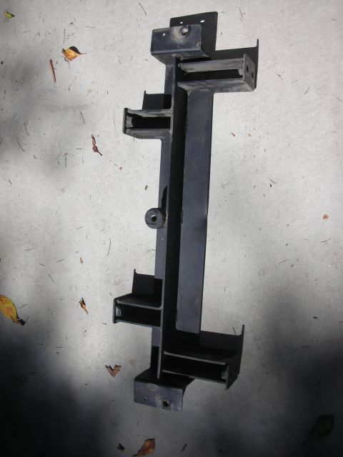 2004 Ford Crown Victoria - A removed black-painted fabricated steel bracket assembly lying on sunlit concrete, built from a long channel spine with several welded squ…