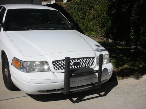 2004 Ford Crown Victoria - Three-quarter front view of a white Ford sedan on a sunlit concrete driveway, wearing a black steel push bumper bolted to the front, black…