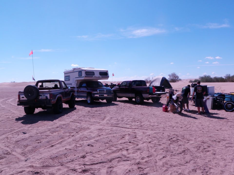 Brown Ford Bronco with spare tire on rear, parked on sand with a camper truck and other vehicles
