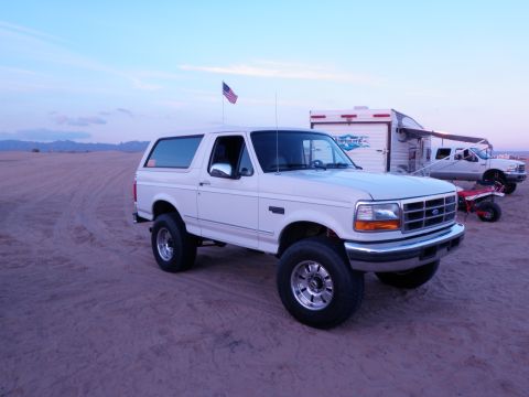 1982 Ford Bronco - White Ford Bronco parked on sand with a trailer and another truck in the background under a dusky sky.