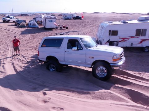 1982 Ford Bronco - A white Ford Bronco is partially submerged in a sand dune, with a campsite and other vehicles in the background.