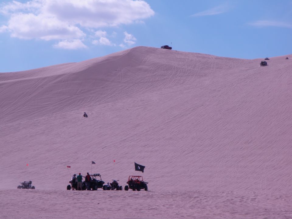 Several off-road vehicles and people gathered on a large sand dune under a blue sky with clouds. Some vehicles are driving on the dune