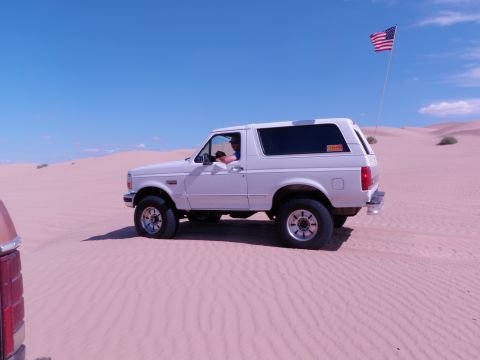 1982 Ford Bronco - White Ford Bronco navigates sand dunes under a clear blue sky, with an American flag flying.