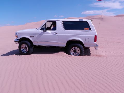 1982 Ford Bronco - A white Ford Bronco is partially buried in pink sand dunes under a clear blue sky with a flag on a pole.