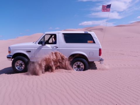 1982 Ford Bronco - A white Ford Bronco is seen driving through sand dunes, kicking up a cloud of dust.