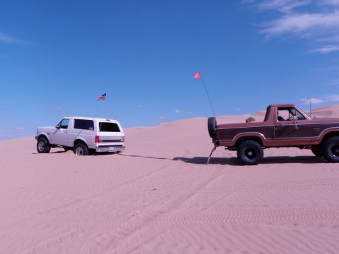 1982 Ford Bronco - A white 1982 Ford Bronco, stuck in the sand, is being towed by a brown pickup truck under a clear blue sky.