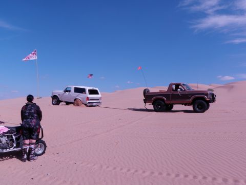 1982 Ford Bronco - A brown pickup truck tows a white SUV out of the sand dunes under a clear blue sky.