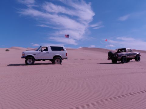 1982 Ford Bronco - Two Ford Bronco SUVs traverse sandy terrain under a bright, cloudy sky.