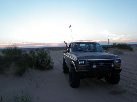 1982 Ford Bronco - A brown pickup truck navigates a sandy track in the desert as the sun sets, with a person waving from the window.