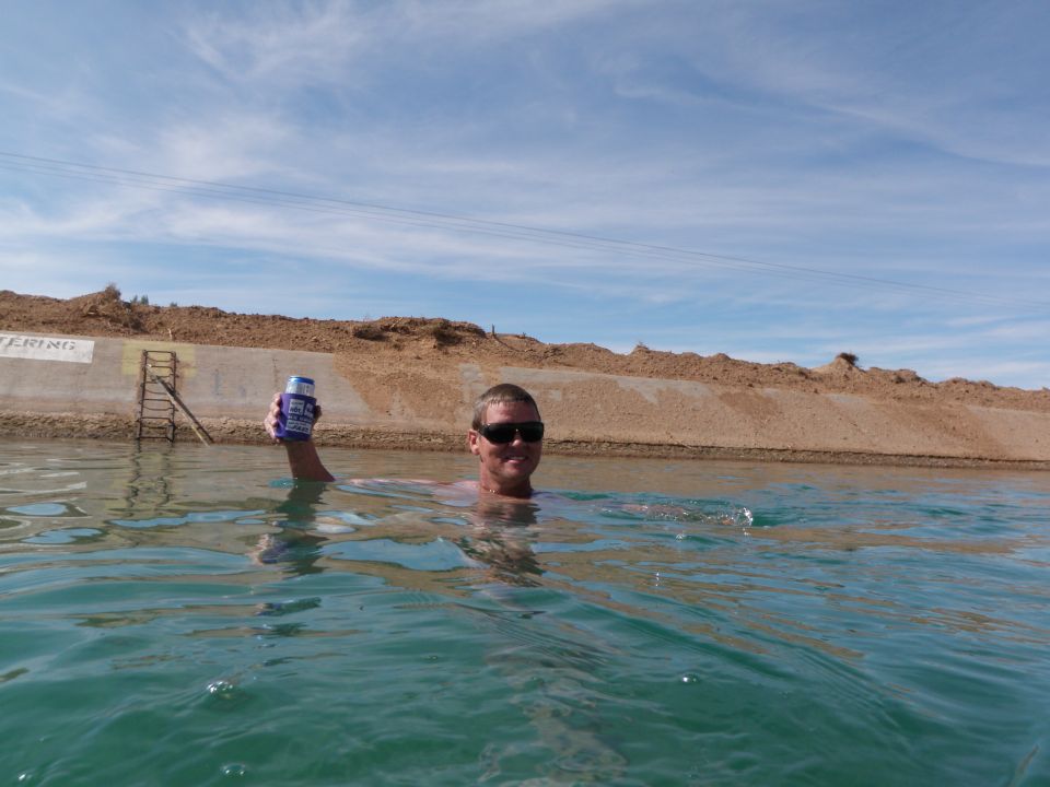 Guy swimming in canal holding a beer can. Dirt embankment and blue sky in background