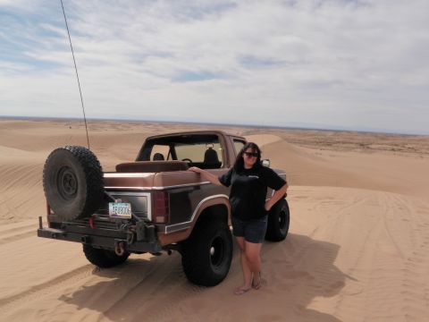 1982 Ford Bronco - A woman stands next to a brown Ford Bronco in a desert landscape with sand dunes under a cloudy sky.