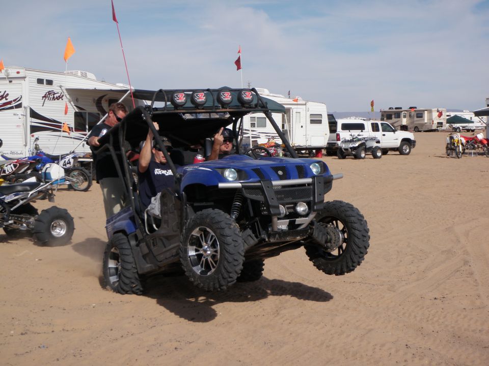 Blue side-by-side off-road vehicle with two people, one giving a middle finger. Other ATVs and RVs in background