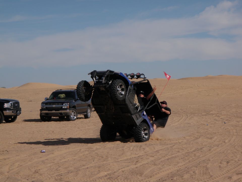 Side-by-side ATV flipping onto its side in the sand with two people trying to hold it up. Another SUV and truck are parked nearby