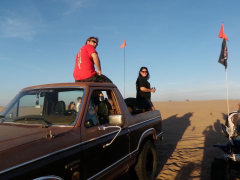 1982 Ford Bronco - Two people sit on the roof of a vintage Ford Bronco in a desert landscape with flags and another vehicle nearby.