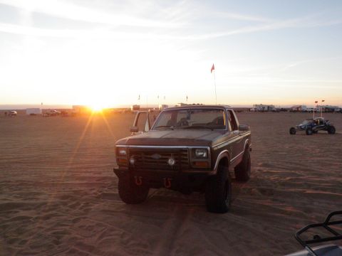 1982 Ford Bronco - A Ford pickup truck sits on the sand as the sun sets over the dunes, with other vehicles visible in the distance.