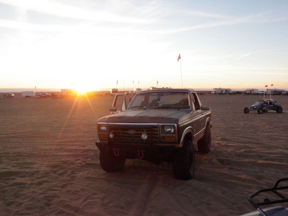 Brown Ford pickup truck parked on sand dunes at sunset, with a dune buggy in the background
