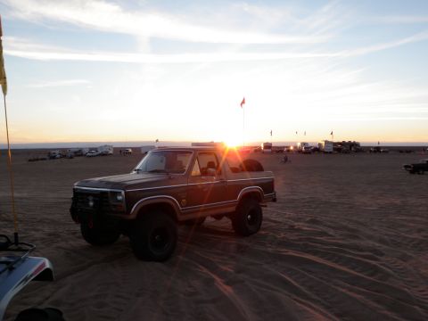 1982 Ford Bronco - A brown Ford Bronco is parked on sand dunes with other vehicles and trailers in the background during sunset.