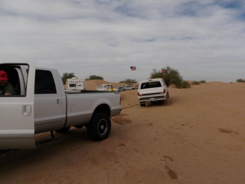 1982 Ford Bronco - A white Ford Bronco is being towed by a pickup truck in a desert area with sand dunes and sparse vegetation.