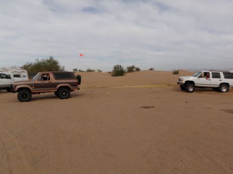 1982 Ford Bronco - Two vehicles, a brown Ford Bronco and a white SUV, are in a sandy desert landscape, with the SUV towing the Bronco.