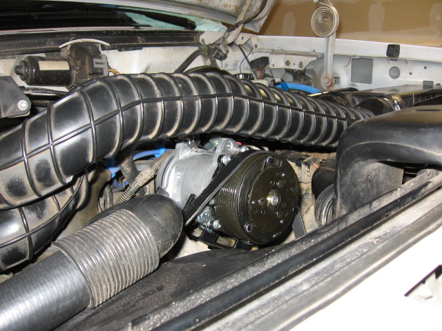 A/C compressor with black clutch pulley in engine bay behind ribbed intake duct