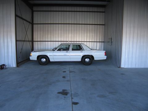 1991 Ford LTD Crown Victoria - A white sedan is parked inside a metal structure, showing its side profile.