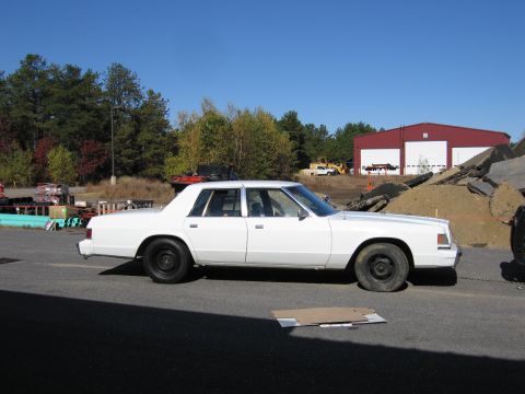 1991 Ford LTD Crown Victoria - A white sedan is seen parked on an asphalt surface, with a red building and construction debris visible in the background.
