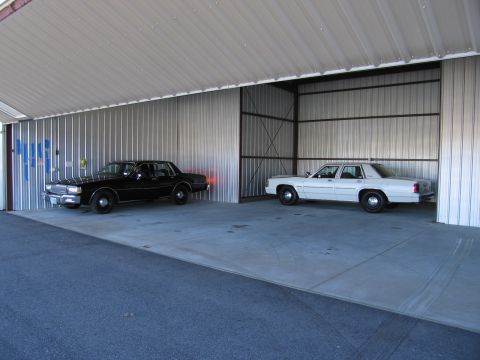 1991 Ford LTD Crown Victoria - Two older model sedans are parked side-by-side under a large metal roofed structure.
