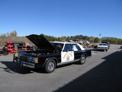 1991 Ford LTD Crown Victoria - A black and white police car with its hood up is parked on asphalt, with other vehicles and debris in the background.