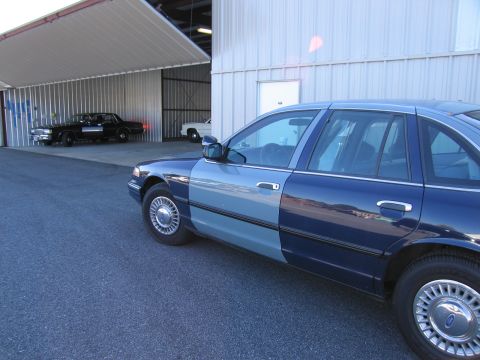 1991 Ford LTD Crown Victoria - A two-tone blue sedan is parked outside a hangar, with two other vehicles visible inside.