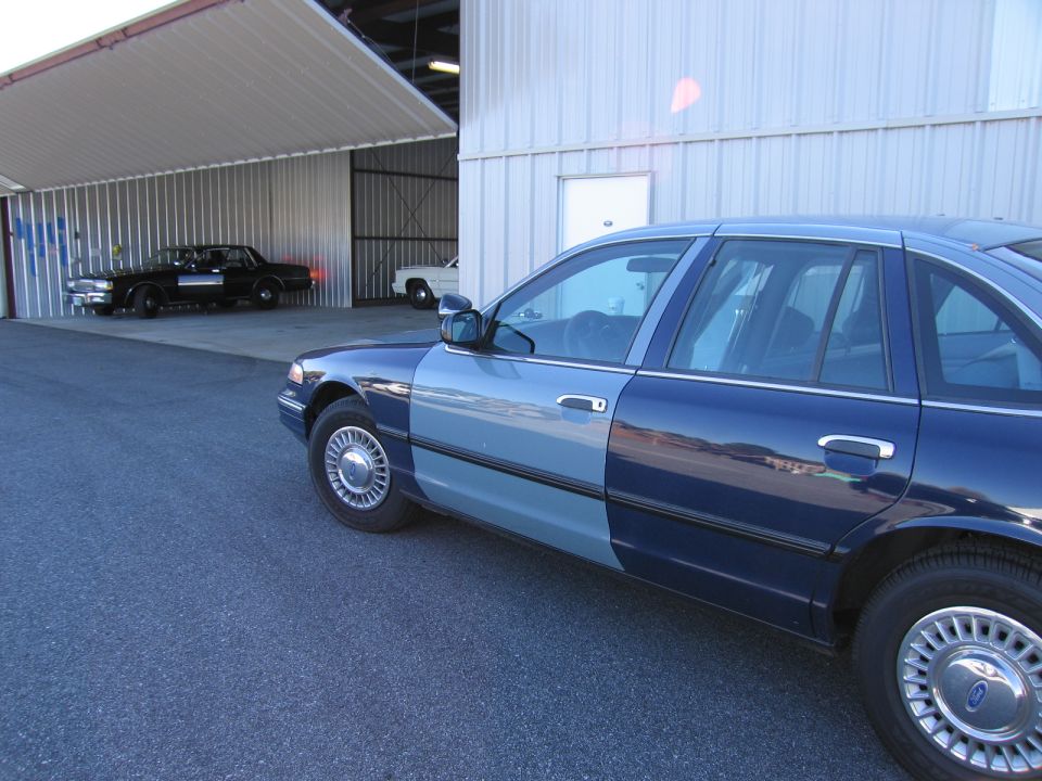 Two-tone blue sedan, 3/4 front view. Two other vehicles are parked in a hangar