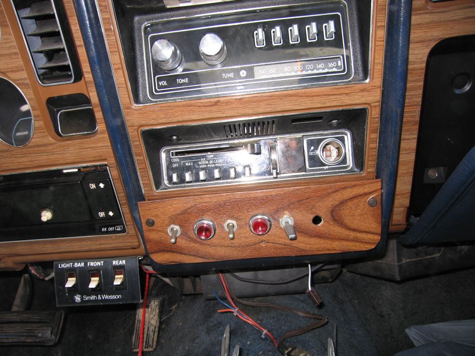 Car dashboard showing a vintage radio, climate controls, and several aftermarket toggle switches and buttons. A Smith & Wesson labeled switch panel is visible