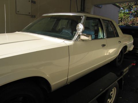1991 Ford LTD Crown Victoria - A white sedan is loaded onto a trailer, showing its side profile and a mounted spotlight.