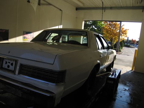 1991 Ford LTD Crown Victoria - A white sedan is parked inside a garage, viewed from the rear, with an open door revealing an autumn scene outside.