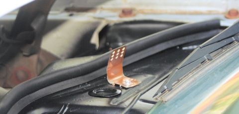 1976 Ford Ranchero - Tight close-up of a small perforated copper-colored ground strap or tab screwed to the black cowl panel at the base of the windshield, next…